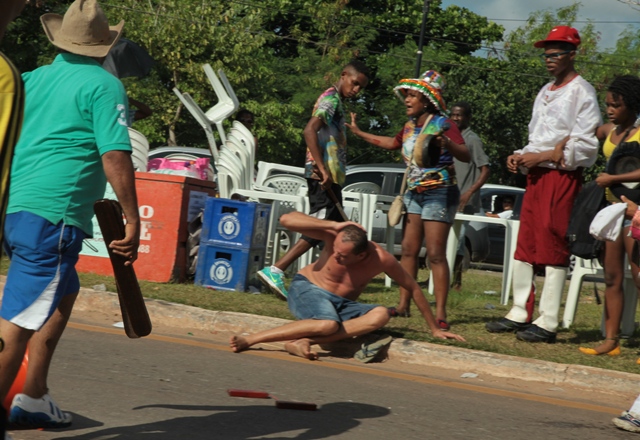 Sentado no meio-fio, homem protege a cabeça, enquanto agressor o ameaça com matraca